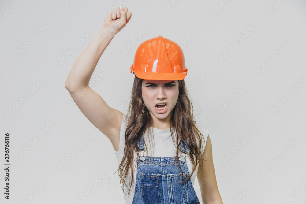 Young pretty girl standing on a white background. During this time wearing a white T-shirt and denim overalls. Dressed in an orange helmet. Young pretty girl standing on a white background. During this time wearing a white T-shirt and denim overalls. Dressed in an orange helmet.