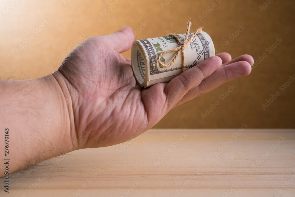 Fototapeta premium A man Hand and a roll of American currency (USD, American dollars) with 100 dollars bank notes on the outside as a symbol of plenty of money on the wooden background.