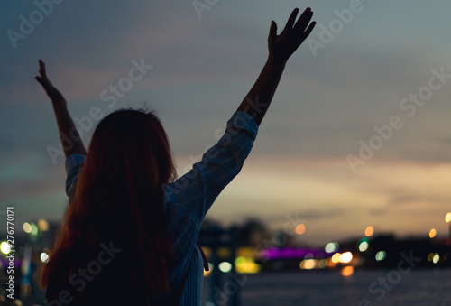 Silhouette behind a young girl raising her hands in the air during golden hour - Woman traveller enjoying watching a beautiful sunset with city bokeh lights - Travel  freedom  lifestyle concept