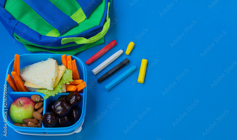 School lunch box with sandwich, nuts, vegetables and fruits on blue background with school supplies. Copy space.