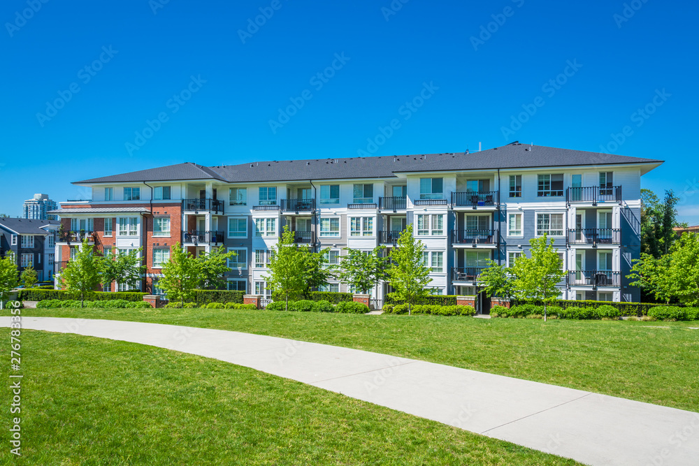 Concrete pathway across green lawn in front of residential condo building. Residential apartment building on sunny day with blue sky. Concrete pathway across green lawn in front of residential condo building. Residential apartment building on sunny day with blue sky.