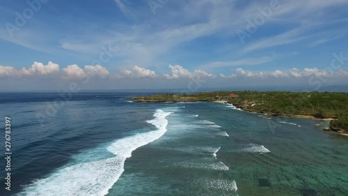 Flying above long foamy ocean waves along tropical island coast line. Village houses and resorts in background. Aerial footage of Nusa Lembongan island.