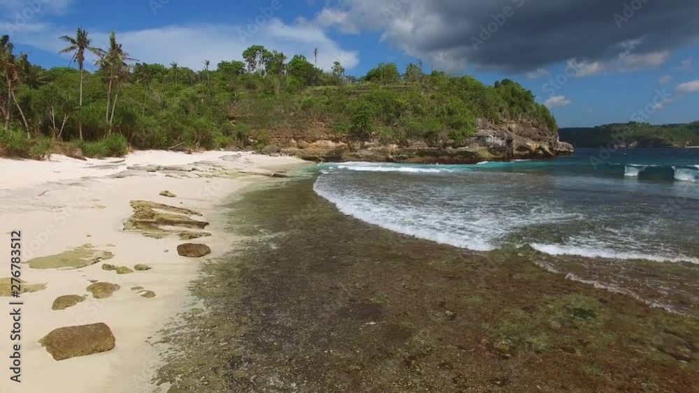 Moving forward along sandy shore of deserted beach on tropical island near the turquoise waters of Indian Ocean towards a low cliff covered with lush green vegetation in sunny weather. Nusa Ceningan. Moving forward along sandy shore of deserted beach on tropical island near the turquoise waters of Indian Ocean towards a low cliff covered with lush green vegetation in sunny weather. Nusa Ceningan.