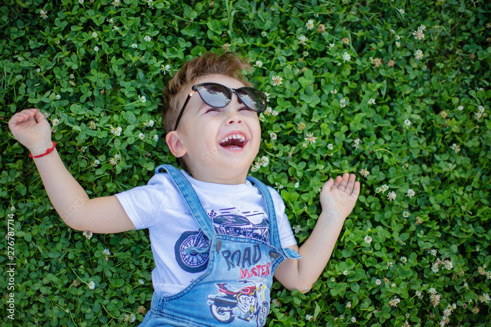 cute little boy in sunglasses laughs lying on the grass in summer in the Park laughs and smiles happily about something dreaming cute little boy in sunglasses laughs lying on the grass in summer in the Park laughs and smiles happily about something dreaming