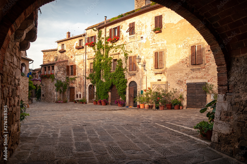 Montemerano, Tuscany. View of the main square of the old medieval town through a red brick arch. Ivy stuck on the wall and ornamental plants. Walls illuminated by the light of the setting sun