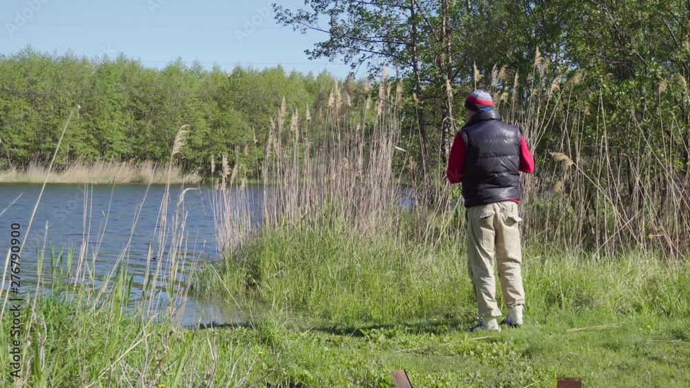 Static shot of fishing. Lone fisherman on the lake fishing with a fishing rod. Sunny morning on the beautiful lakeside. On the shore, high vegetation. Static shot of fishing. Lone fisherman on the lake fishing with a fishing rod. Sunny morning on the beautiful lakeside. On the shore, high vegetation.