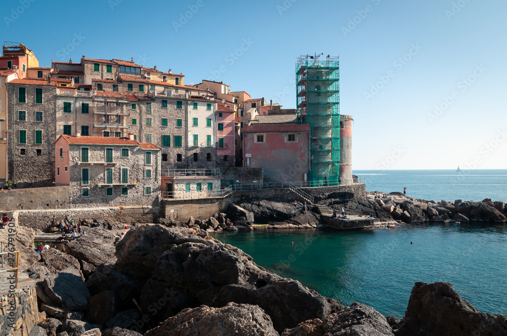 Tellaro, La Spezia, Italy. Typical foreshortening of the ancient village: agglomeration of stone houses with pastel colors. Church of San Giorgio near the sea. Rocks in the foreground