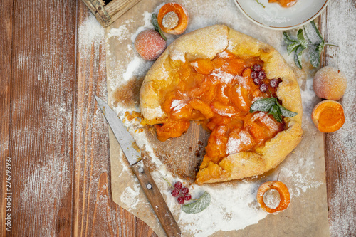 Homemade fruit pie (galette) made with fresh organic apricotes with powdered sugar on wooden table, top view. Plate with a cut piece of cake. Open pie, apricot tart on parchment paper. photo