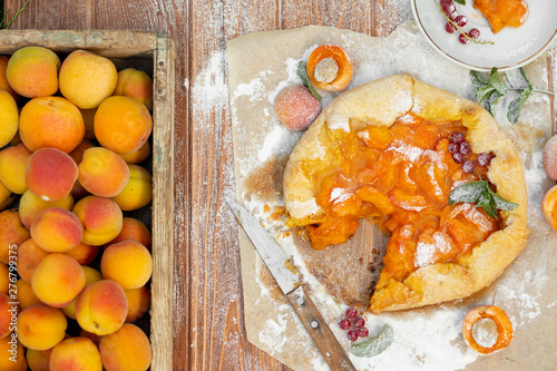 Homemade fruit pie (galette) made with fresh organic apricotes with powdered sugar on wooden table, top view. Plate with a cut piece of cake. Open pie, apricot tart on parchment paper. photo