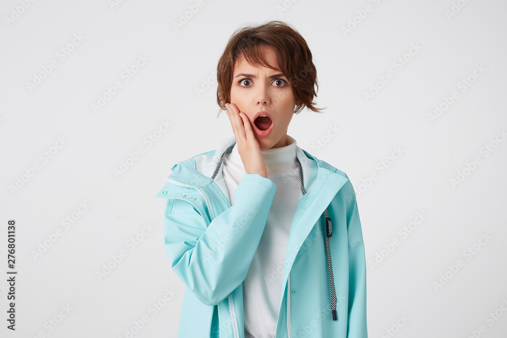 Shocked short-haired young woman in white golf and blue rain coat, stands over white background with wide open mouth and surprised expression, touches cheek, looks at the camera with wide open eyes. Shocked short-haired young woman in white golf and blue rain coat, stands over white background with wide open mouth and surprised expression, touches cheek, looks at the camera with wide open eyes.