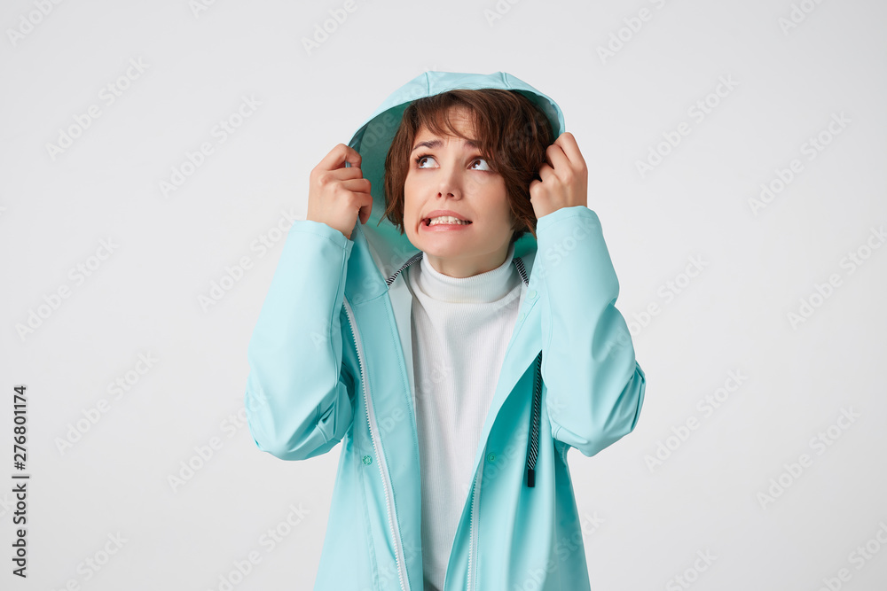 Cute disgusting short-haired curly woman in white golf and light blue rain coat, hiding under the hood from rain and looking up at the left side, stands over white background. Cute disgusting short-haired curly woman in white golf and light blue rain coat, hiding under the hood from rain and looking up at the left side, stands over white background.