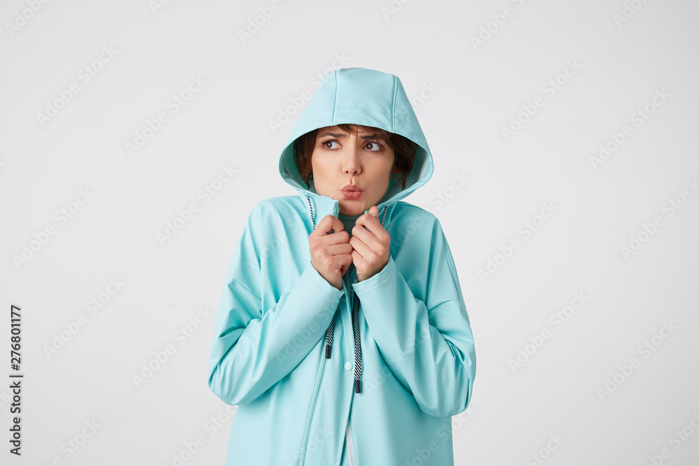 Cute frozen short-haired curly woman in white golf and light blue rain coat, hiding under the hood from rain and looking away, stands over white background. Cute frozen short-haired curly woman in white golf and light blue rain coat, hiding under the hood from rain and looking away, stands over white background.