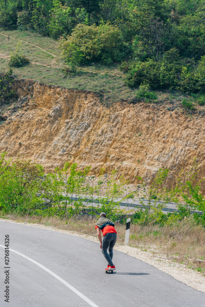 Male caucasian longboarder riding downhill on an empty road doing a speed tuck and grabbing the board while driving the longboard fast. Wearing a red t-shirt green hat and black jeans.