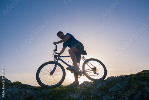 Strong fit male mountain biker performing stunts on rocky terrain on a sunset while wearing a blue shirt and riding a blue bike © qunica.com
