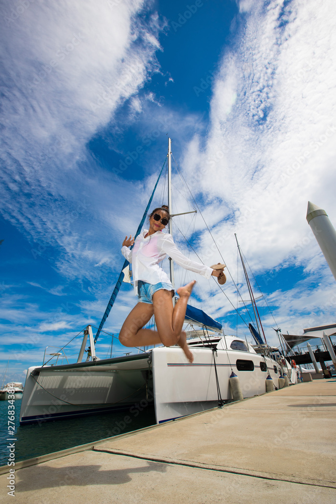 Beautiful Mix Race Tanned skin Woman high jump along Luxury Yachts in Marina Bay Club, white shirt Jean Shirt Girl post as Fashion Model in docking pier under summer blue sky in vacation holiday