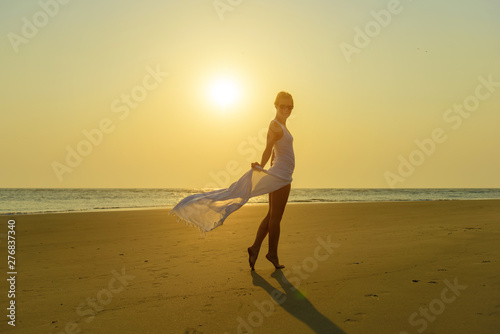 Attractive stylish blonde in sunglasses and light white dress stands barefoot on yellow sand on background of sea and setting sun. Girl on the beach on hot summer evening against beautiful landscape.