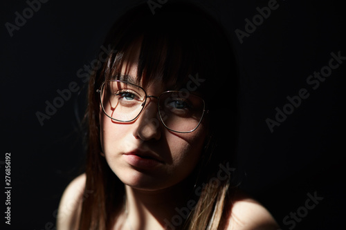Close up sensual portrait of young blue eyed Caucasian female with dark hair and charming appearance, sitting in dark room under sunny ray, wearing big elegant eye glasses, looking at camera deeply.