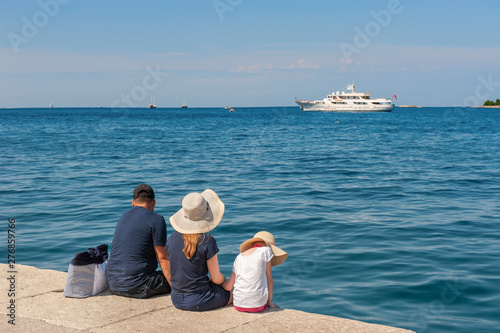 rear view of the family sitting on a stone pier on a summer day. The family admires the beautiful sea view from the shore