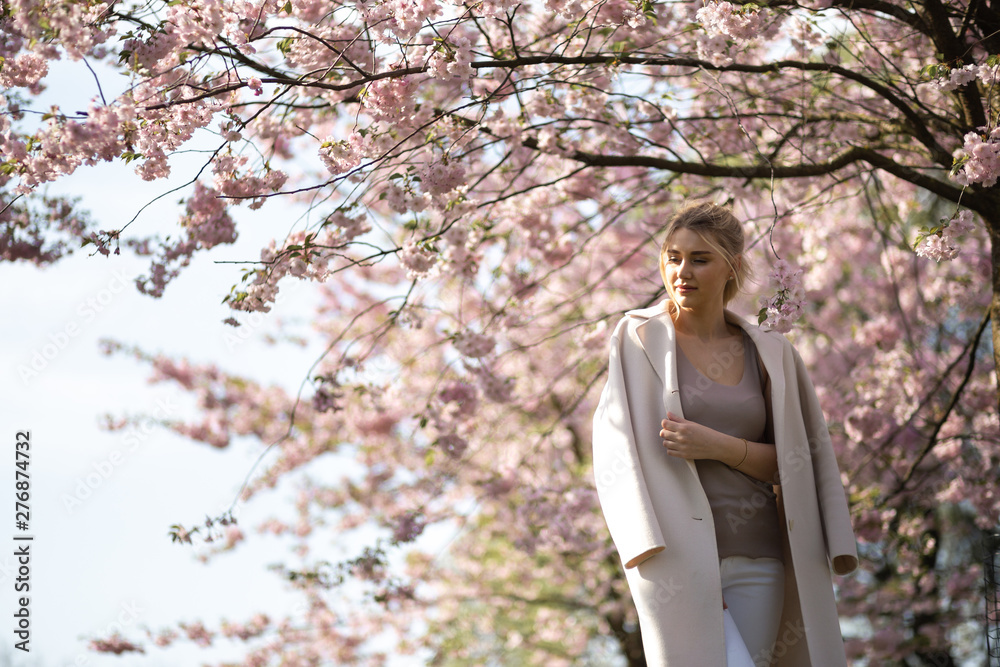 Beautiful blonde young woman in Sakura Cherry Blossom park in Spring enjoying nature and free time during her traveling tourist free time - Wearing white pants and t-shirt with a beige jacket