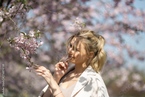 Beautiful blonde young woman in Sakura Cherry Blossom park in Spring enjoying nature and free time during her traveling tourist free time - Wearing white pants and t-shirt with a beige jacket