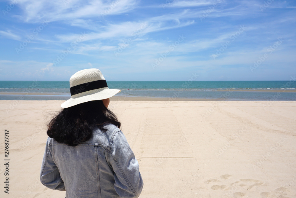 Asian woman wearing hat and jeans jacket standing at the beautiful beach looking at the sea