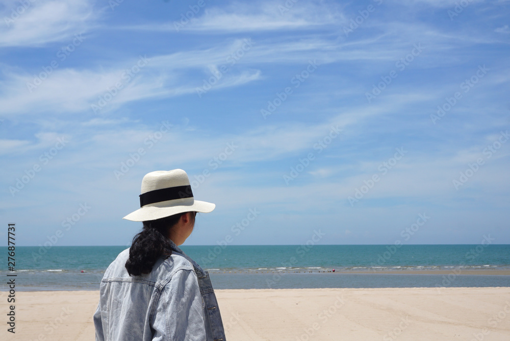 Asian woman wearing hat and jeans jacket standing at the beautiful beach looking at the sea