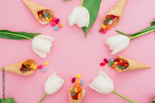 Fototapeta Naklejka Na Ścianę i Meble -  Frame made of sugar candy in waffle cones and white tulip flowers on pink background. Flat lay