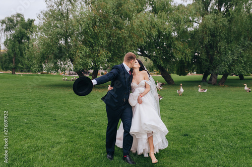 Smiling and cheerful newlyweds stroll and kiss against the background of green grass in the park. Wedding portrait of a stylish groom with a hat and a beautiful bride with curly hair and white dress. photo