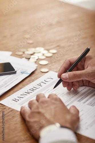 cropped view of businessman in suit filling in bankruptcy form at wooden table with coins