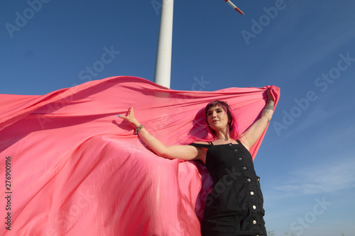 Pink-haired young woman with pieces of pink cloth dance near the field with wind generators . Windmill on sunset. Fashion portrait