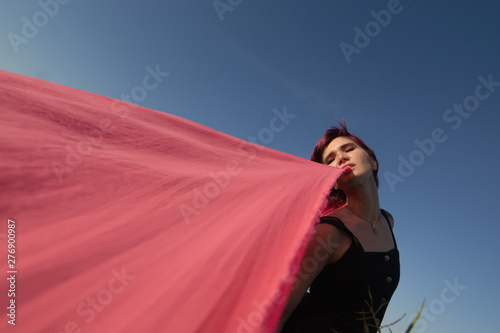 Pink-haired young woman with pieces of pink cloth dance near the field with wind generators . Windmill on sunset. Fashion portrait