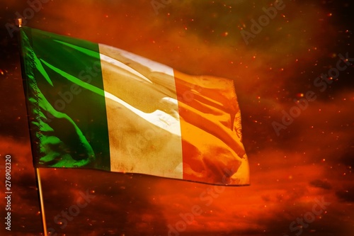 Fotografie Fluttering Ireland flag on crimson red sky with smoke pillars background