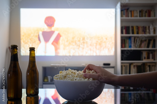 Home cinema entertainment: watching a film from a video projector in a room. Dim living room with a cinematic picture projected on the wall, human hand grabbing popcorn