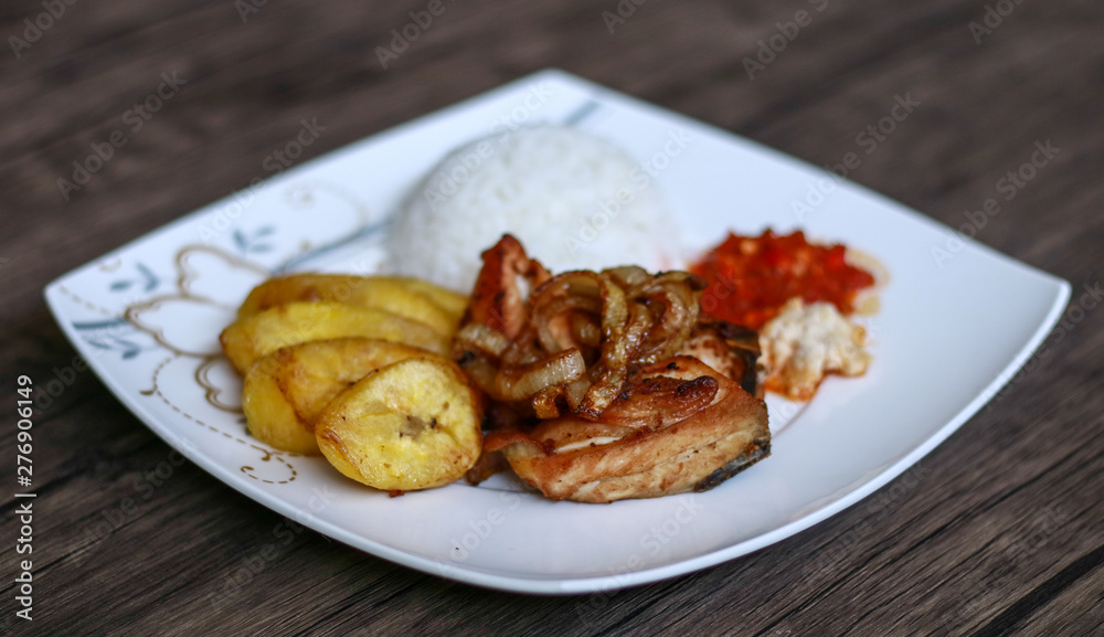 African fried chicken with fried bananas, rice, fried onions, chili sauce and candlenut sauce. Wood background. African fried chicken with fried bananas, rice, fried onions, chili sauce and candlenut sauce. Wood background.