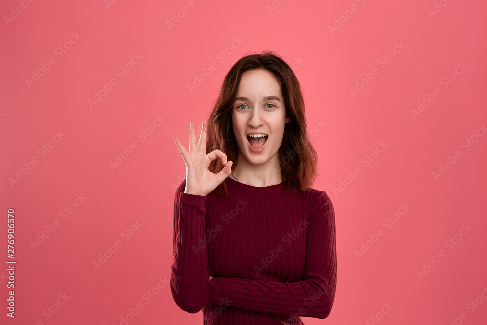 Happy young lady standing isolated over dark pink background looks at the camera while holding palm of a hand near her eye showing gesture of OK-sign. Happy young lady standing isolated over dark pink background looks at the camera while holding palm of a hand near her eye showing gesture of OK-sign.