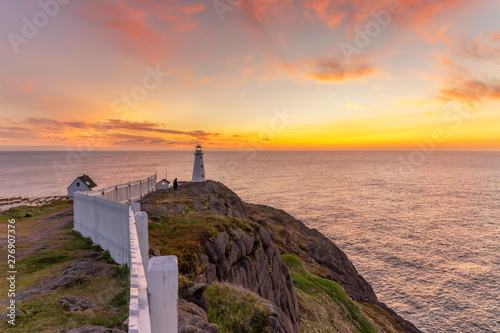 Soft pink and orange clouds light up the sky before sunrise over a white lighthouse sitting at the edge of a rocky cliff. Cape Spear National Historic Site, St Johns Newfoundland. 