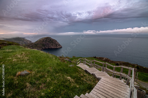 A wooden staircase leading down to a dramatic coastal seascape with ominous clouds and fog rolling in. An iceberg in the ocean off shore. Newfoundland Canada.   photo