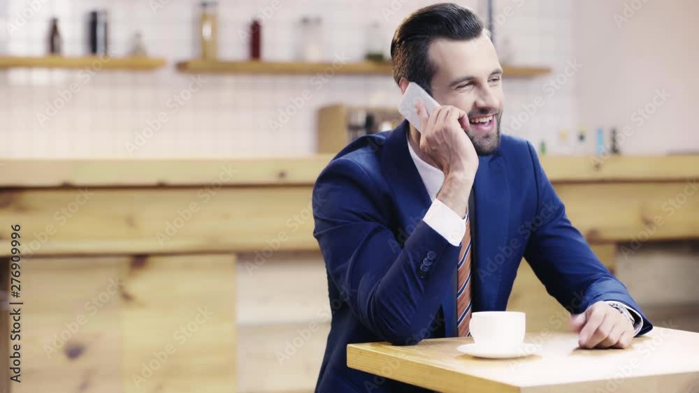 handsome and smiling businessman in suit talking on smartphone, looking at watch in cafe handsome and smiling businessman in suit talking on smartphone, looking at watch in cafe