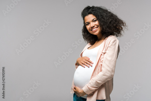 cheerful young pregnant african american woman touching belly on grey photo