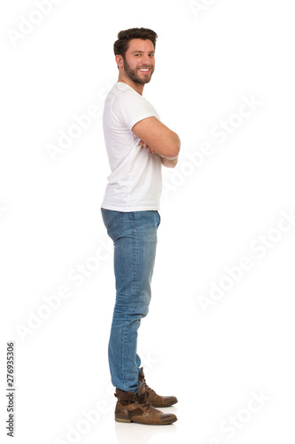 Side View Of Young Man In Jeans And White T-shirt Is Standing With Arms Crossed, Looking At Camera