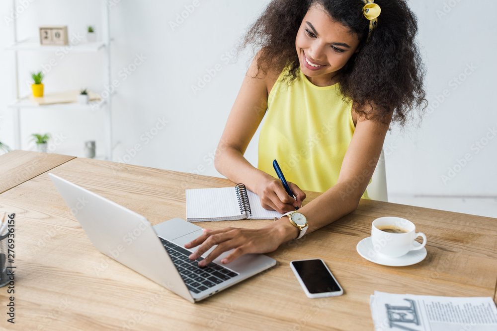 happy african american woman holding pen near notebook and looking at laptop happy african american woman holding pen near notebook and looking at laptop
