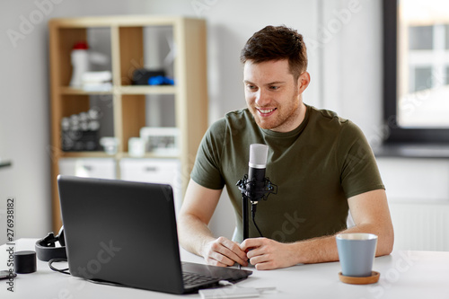 technology, mass media and podcast concept - happy young male audio blogger with laptop computer and microphone broadcasting at home office