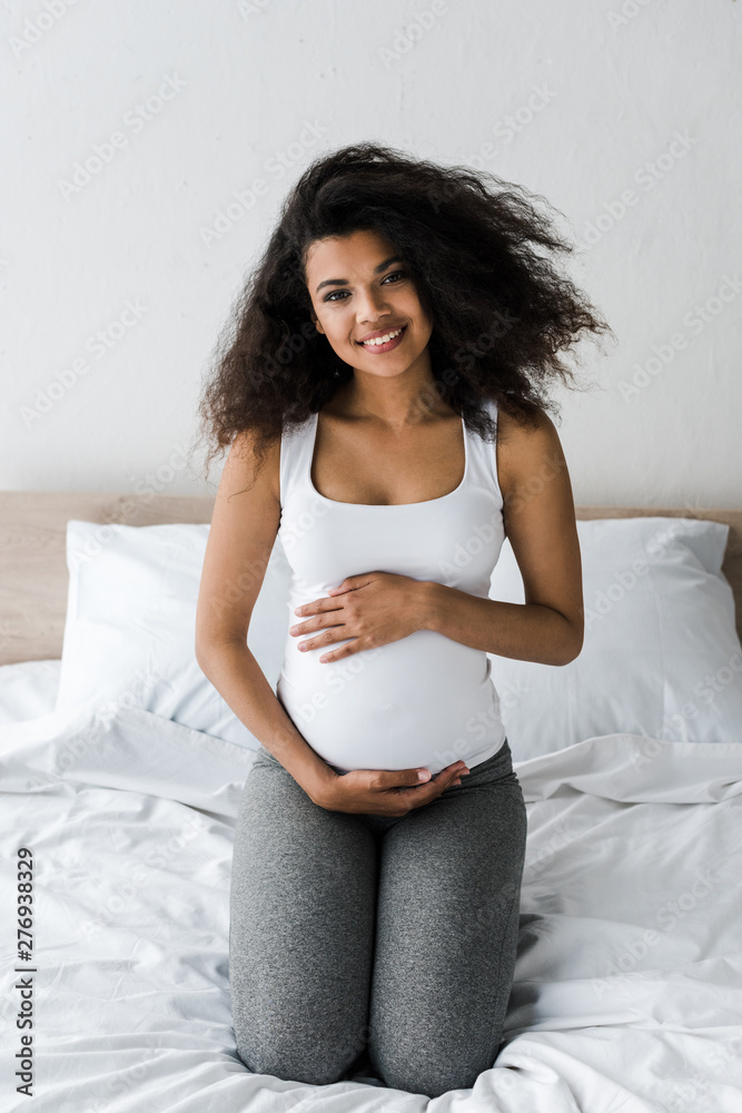 happy curly african american pregnant woman touching belly while sitting on bed happy curly african american pregnant woman touching belly while sitting on bed