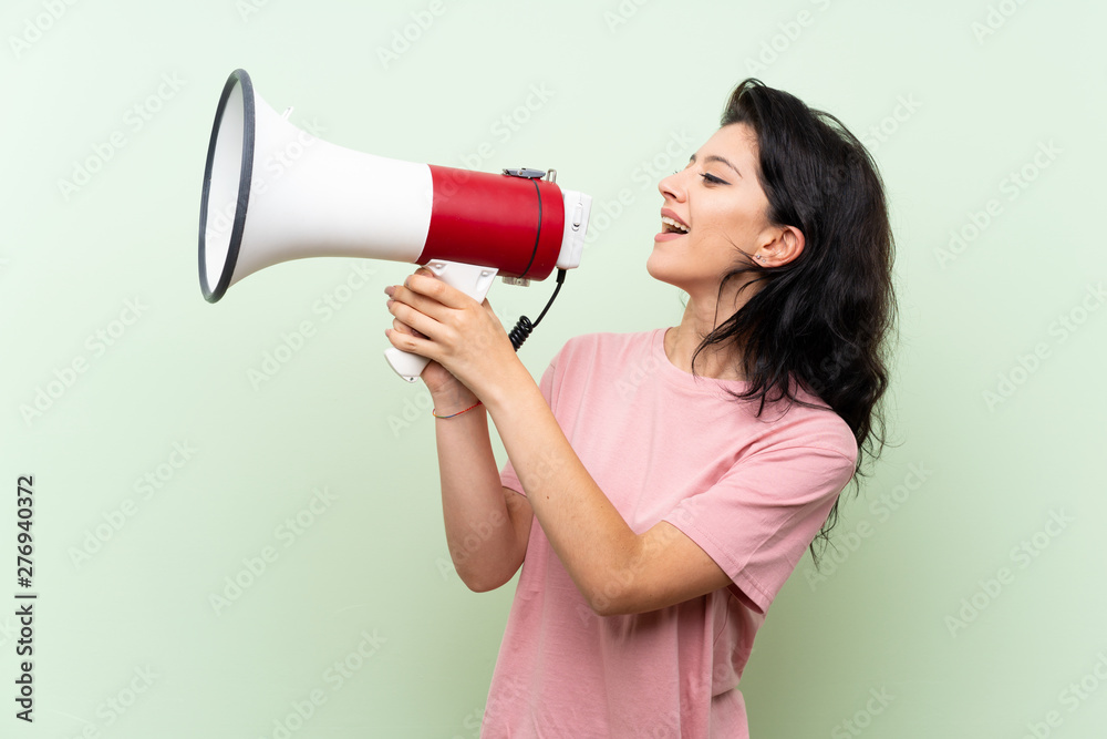 Young woman over isolated green background shouting through a megaphone Young woman over isolated green background shouting through a megaphone