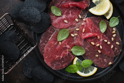 Above view of carpaccio beef with green basil, lemon, pine nuts and black potato chips, close-up