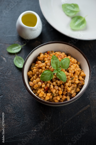 Bowl of fregola pasta with fresh green basil and olive oil, studio shot on a dark brown stone background with space