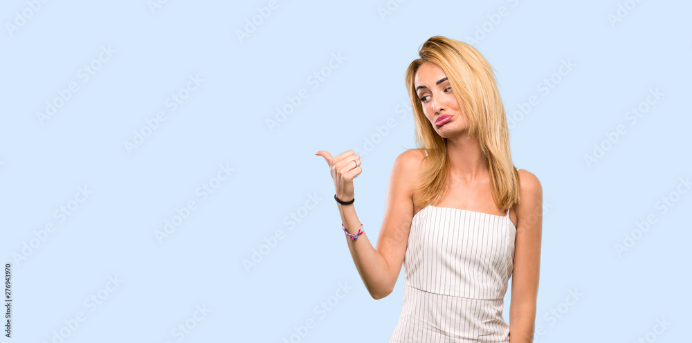Young blonde woman unhappy and pointing to the side over isolated blue background Young blonde woman unhappy and pointing to the side over isolated blue background