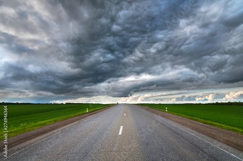 Evening landscape of suburban asphalt road with low textured dramatic clouds and green fields on the sides
