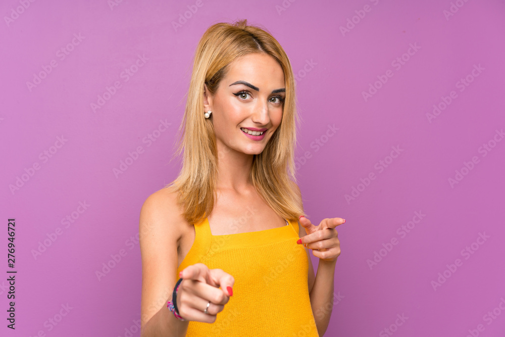 Young blonde woman over purple background points finger at you Young blonde woman over purple background points finger at you