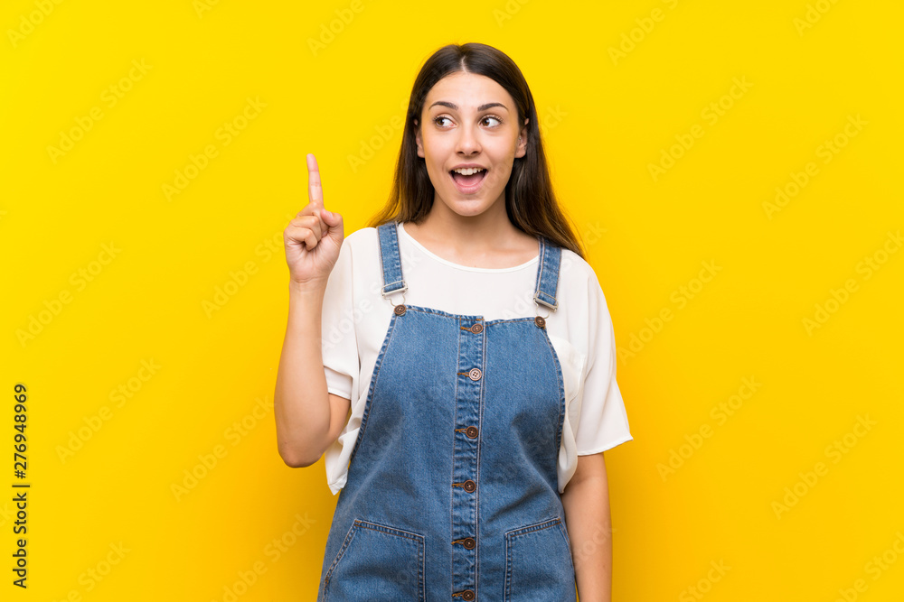 Young woman in dungarees over isolated yellow background intending to realizes the solution while lifting a finger up Young woman in dungarees over isolated yellow background intending to realizes the solution while lifting a finger up